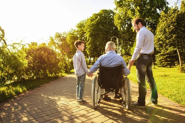 O velho em uma cadeira de rodas, seu filho e neto estão andando no parque — Fotografia de Stock
