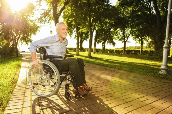 El viejo está sentado en una silla de ruedas en el parque y tratando de ir —  Fotos de Stock