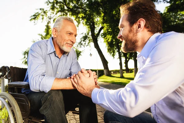 De oude man in een rolstoel met zijn zoon in het park. Theyre wandelen. Een man houdt een oude man bij de hand — Stockfoto