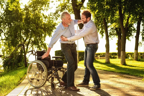 El viejo intenta salir de la silla de ruedas. en muletas. Su hijo lo ayuda. —  Fotos de Stock