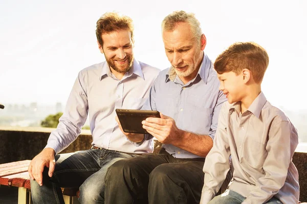 El anciano su hijo y su nieto están sentados en el banco viendo algo en la tableta — Foto de Stock