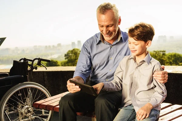 El viejo le está mostrando algo a su nieto en la tableta. Muy cerca está la silla de ruedas viejo — Foto de Stock