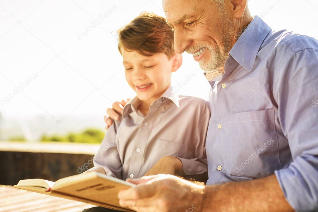 An old man shows something to his grandson in a book. They are sitting on a bench