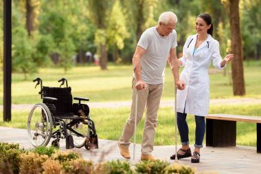 Doktor hasta koltuk değneği üzerinde yürümek için yardımcı olur. Tekerlekli sandalye geride