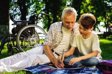 Yaşlı adam ve torunu pikniğe oturuyor. Onlar bir defter kalem ile çizin. Tekerlekli sandalye