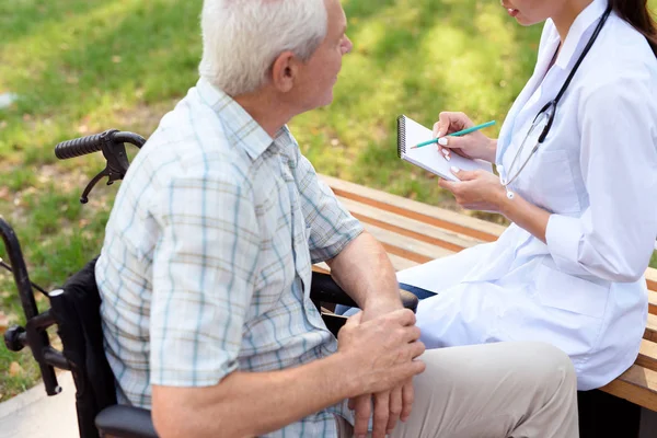 O velho em uma cadeira de rodas comunicando com o médico no parque de verão. Doutor segura um caderno em sua mão — Fotografia de Stock