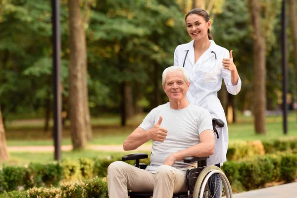 O médico e o paciente que está sentado na cadeira de rodas seguram os polegares para cima e sorriem — Fotografia de Stock