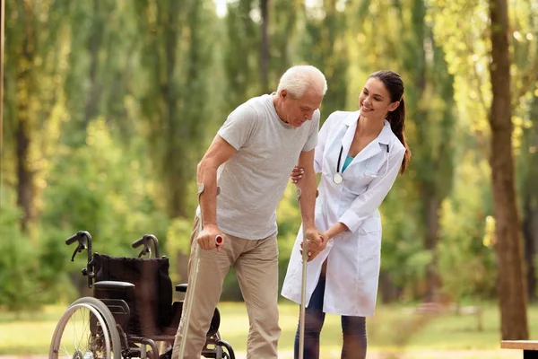 El médico ayuda al anciano a pararse sobre muletas. Silla de ruedas dejada atrás —  Fotos de Stock