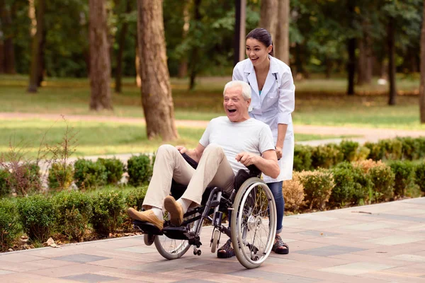 Läkaren rullar en äldre patient på en rullstol i parken. De har roligt — Stockfoto