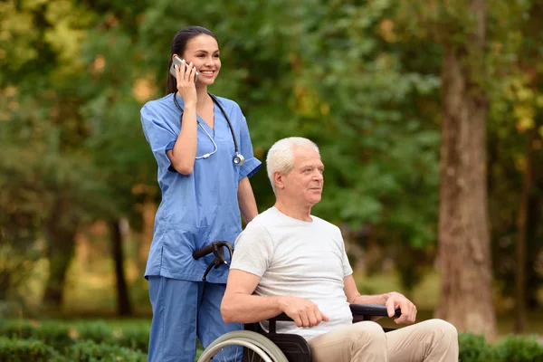 Sjuksköterska och gubbe som sitter i rullstol i parken. Sjuksköterska talar på mobiltelefon — Stockfoto