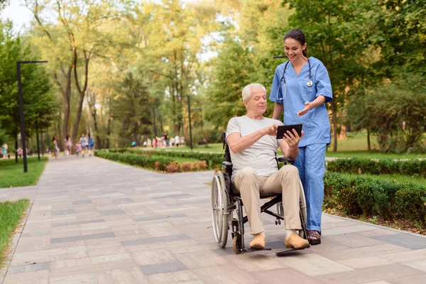 Um homem velho sentado em uma cadeira de rodas mostra à enfermeira algo em seu tablet — Fotografia de Stock
