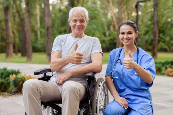 Uma enfermeira e um velho que se senta em uma cadeira de rodas estão mostrando um polegar para cima — Fotografia de Stock