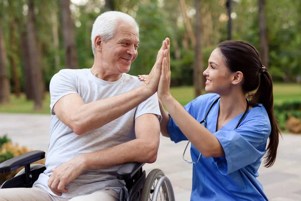 En sjuksköterska och en gammal man i rullstol hög fem i en sommar park — Stockfoto