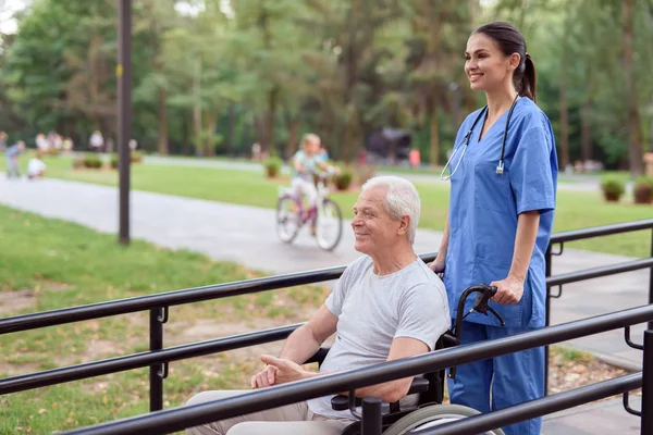 En gammal man i en rullstol och en sjuksköterska promenera längs en brygga i parken — Stockfoto
