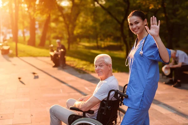Una enfermera está saludando a alguien. Detrás de ella en la silla de ruedas se sienta un anciano y sonríe — Foto de Stock