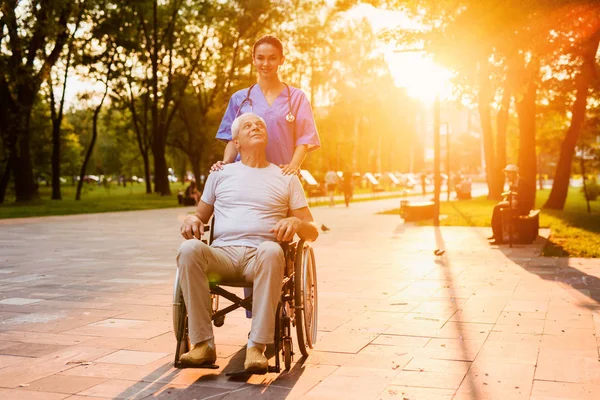 Un anciano que se sienta en una silla de ruedas y una enfermera en el parque al atardecer —  Fotos de Stock