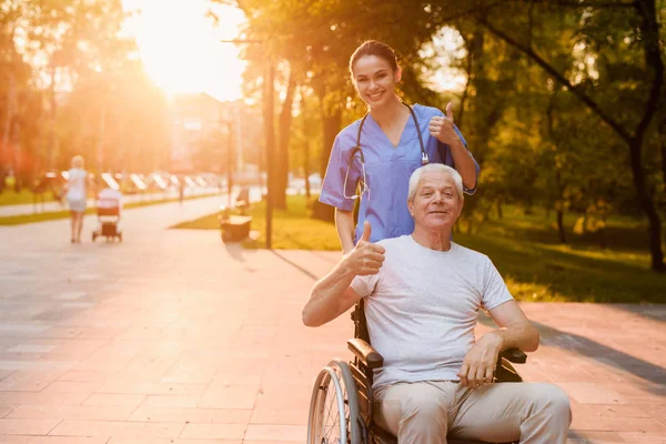 Enfermera y anciano que se sienta en una silla de ruedas mostrando un pulgar hacia arriba en el parque al atardecer —  Fotos de Stock