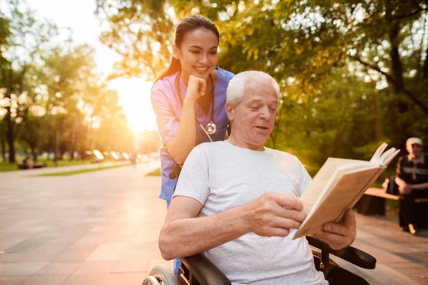 El viejo que está sentado en una silla de ruedas está leyendo un libro. Una enfermera detrás del anciano y también mirando el libro — Foto de Stock