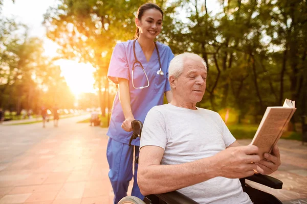 Um velho que se senta em uma cadeira de rodas e uma enfermeira que está atrás dele lendo um livro no parque ao pôr do sol — Fotografia de Stock