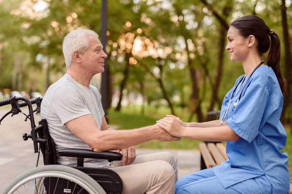 En gubbe som sitter i rullstol och en sjuksköterska som sitter bredvid bänken håller händerna — Stockfoto