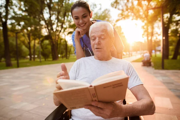 Uma enfermeira observa enquanto o velho lê um livro no parque ao pôr-do-sol — Fotografia de Stock