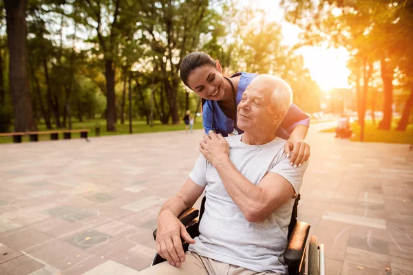 Старик, сидящий в инвалидном кресле, держит медсестру за руку и кладет руку ему на плечо. — стоковое фото