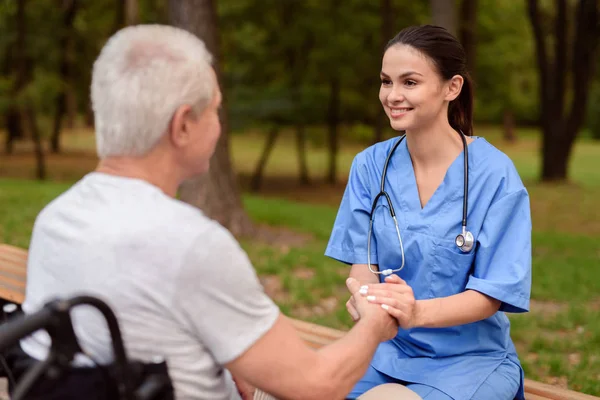 Zdravotní sestra se usmívá a drží ruku starého muže, který sedí vedle vozíku v parku — Stock fotografie