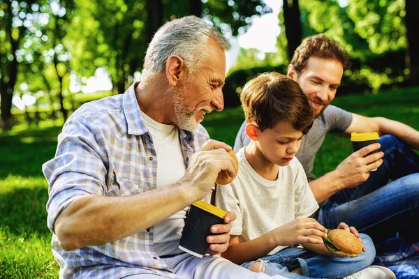 Чоловік, його старший батько і син сидять у парку на пікніку. Хлопчик сидить зі смартфоном у руках — стокове фото