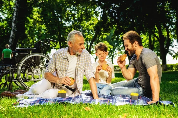 Un hombre, su anciano padre e hijo están sentados en un parque en un picnic. Un niño está sentado con un teléfono inteligente en sus manos — Foto de Stock