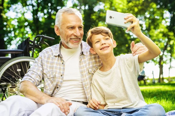 Feliz anciano haciendo selfie con su nieto sentado en el parque en el picnic —  Fotos de Stock