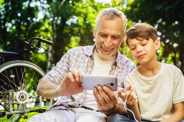 Le vieux et son petit-fils sont assis sur un pique-nique et regardent quelque chose sur le smartphone — Photo