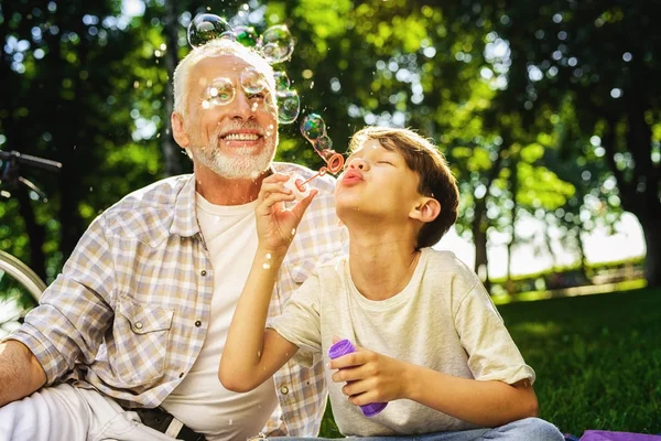 The old man and grandson are sitting on a picnic. The boy blows bubbles — Stock Photo, Image