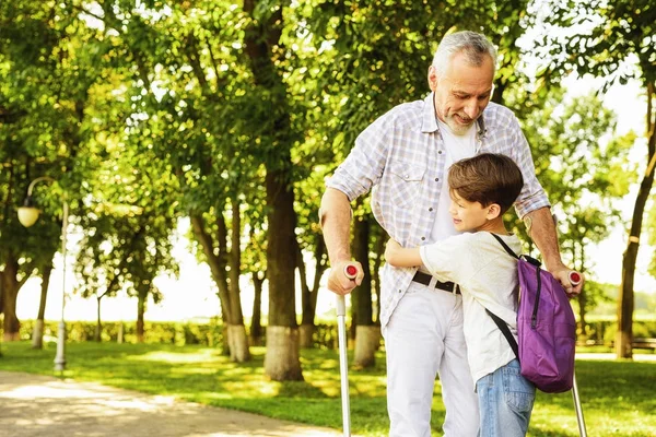 En pojke och en gubbe på kryckor promenader i parken. Pojken håller gammalt mans hand — Stockfoto