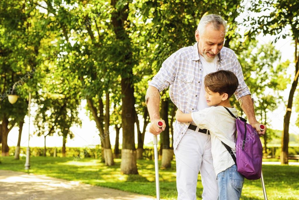 A boy and an old man on crutches are walking in the park. The boy is holding the old mans hand