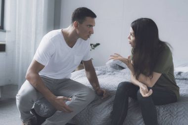 Evli bir çift yatak odasında gri bir yatağın kenarında oturan ve birbirleri ile kavga