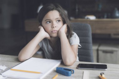 Esmer kız sıkılmış bir ifade ile masasında oturuyor. Yanındaki beyaz bir defter ve onun smartphone olduğunu