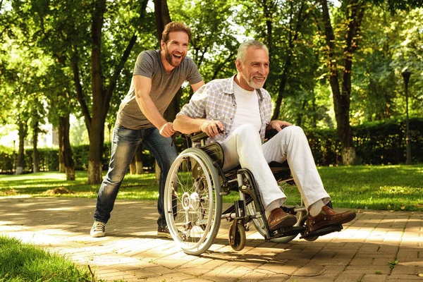 Συνταξιούχος σε μια αναπηρική καρέκλα και ο ενήλικος γιος είναι το περπάτημα γύρω από το πάρκο. Είναι χαρούμενος και να διασκεδάσουν — Φωτογραφία Αρχείου