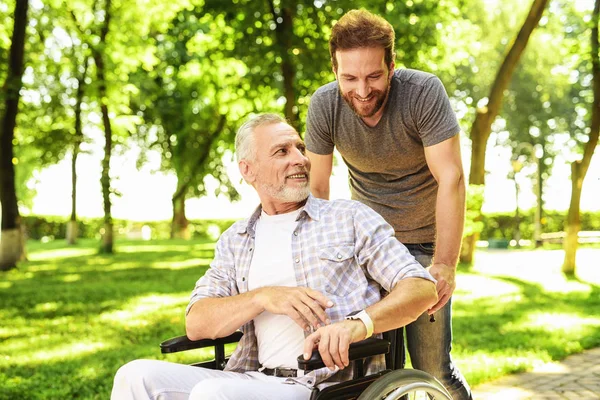 Een man en zijn bejaarde vader lopen in het park. Een man draagt zijn vader in een rolstoel — Stockfoto