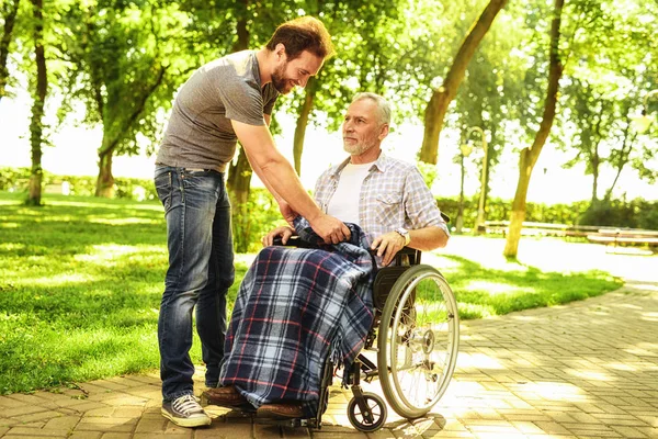 En man och en gammal man i en rullstol i parken. Mannen omfattas den gamla mannen med en matta — Stockfoto