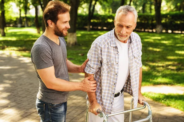 El anciano camina por el parque en caminantes para adultos. Su hijo lo ayuda. — Foto de Stock