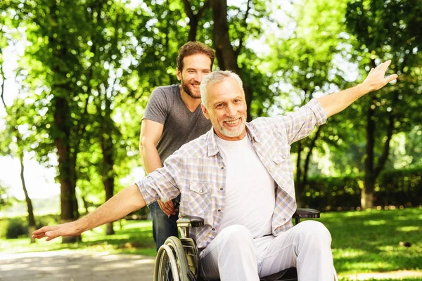 El anciano en silla de ruedas está caminando por el parque con su hijo adulto. Hacen el tonto. —  Fotos de Stock