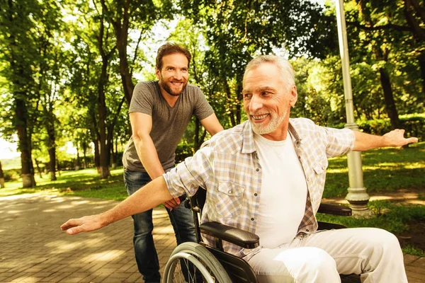 O velho em uma cadeira de rodas está andando no parque com seu filho adulto. Eles brincam. — Fotografia de Stock