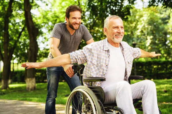 De oude man in een rolstoel is wandelen in het park met zijn volwassen zoon. Ze gek rond — Stockfoto