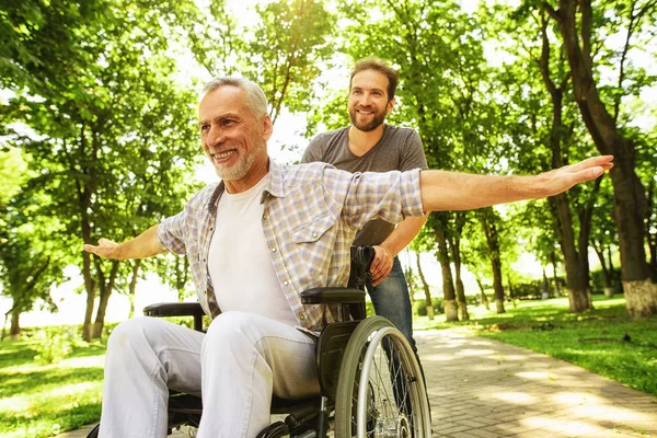 The old man on a wheelchair is walking in the park with his adult son. They fool around — Stock Photo, Image