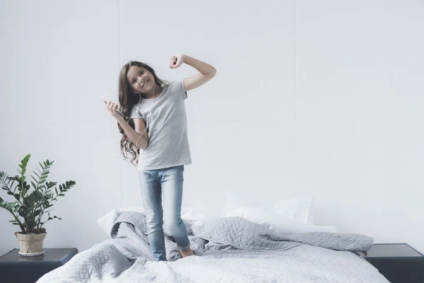 Küçük bir siyah saçlı kız yatakta bir smartphone içinde elleri ve kulaklık ile dans ediyor — Stok fotoğraf