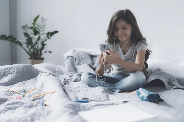 Uma menina ouve sua música favorita em um smartphone cinza com a ajuda de um par de fones de ouvido e sorrisos — Fotografia de Stock