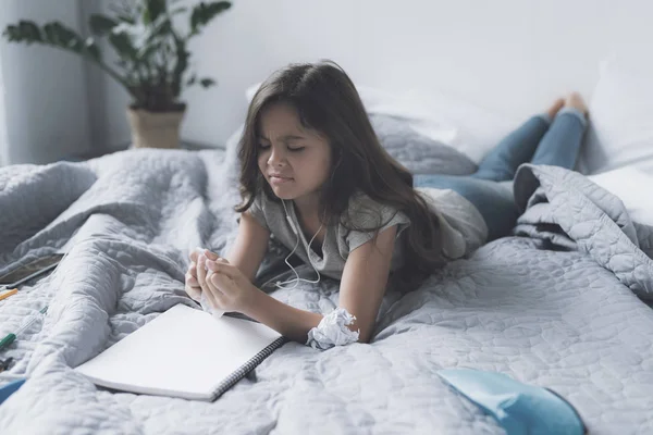 Маленькая темноволосая девочка лежит на кровати в белых наушниках, слушает музыку, смотрит на белую тетрадь и хмурится. — стоковое фото