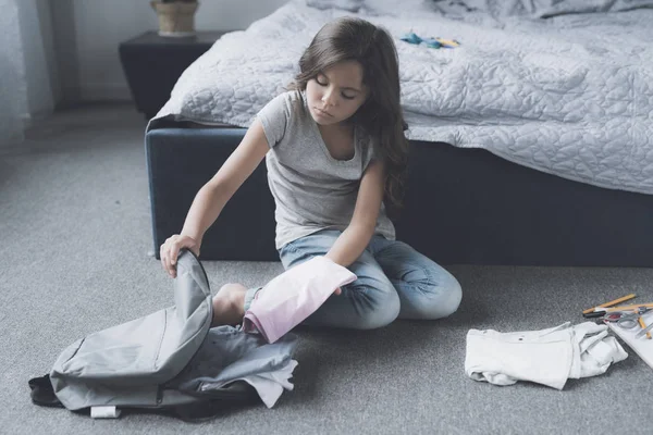 Черноволосая девушка сидит на полу и забирает свою одежду в сером рюкзаке, чтобы сбежать из дома. — стоковое фото