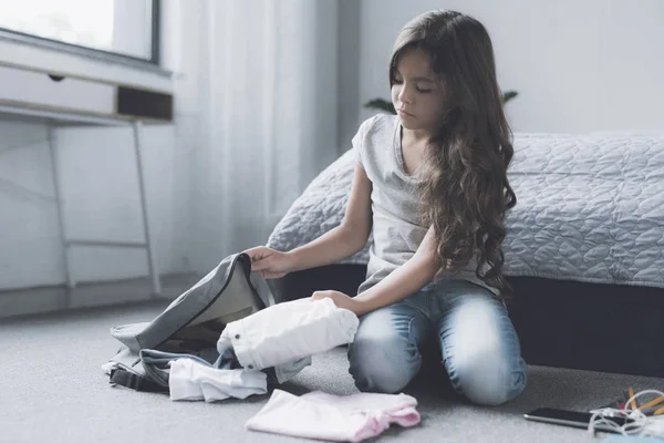 Грустная темноволосая девушка сидит на полу рядом с кроватью и собирает вещи в сером рюкзаке. — стоковое фото