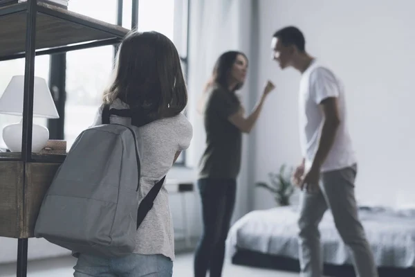 Pendant que les parents grondent dans la chambre, leur fille avec un sac à dos derrière elle se tient à côté des étagères — Photo